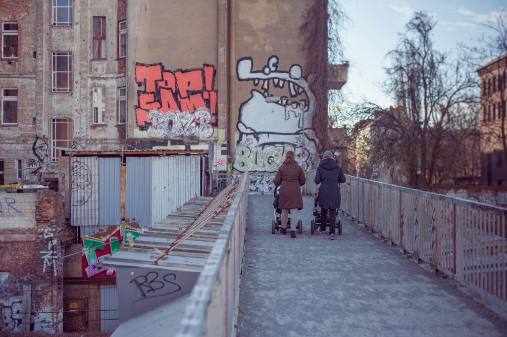 Mothers walking with strollers on graffiti-lined urban street bridge.