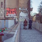 Mothers walking with strollers on graffiti-lined urban street bridge.
