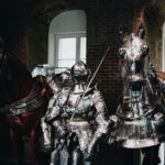 Medieval knight in full armor with a swordsman and armored horse indoors.