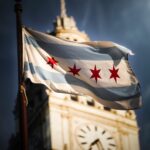 Chicago flag prominently waves in the foreground with the iconic clock tower blurred in the background.