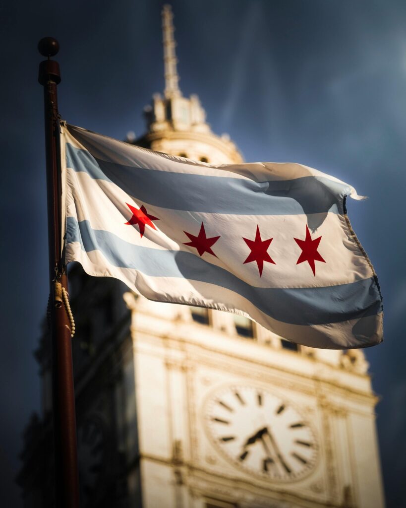 Chicago flag prominently waves in the foreground with the iconic clock tower blurred in the background.