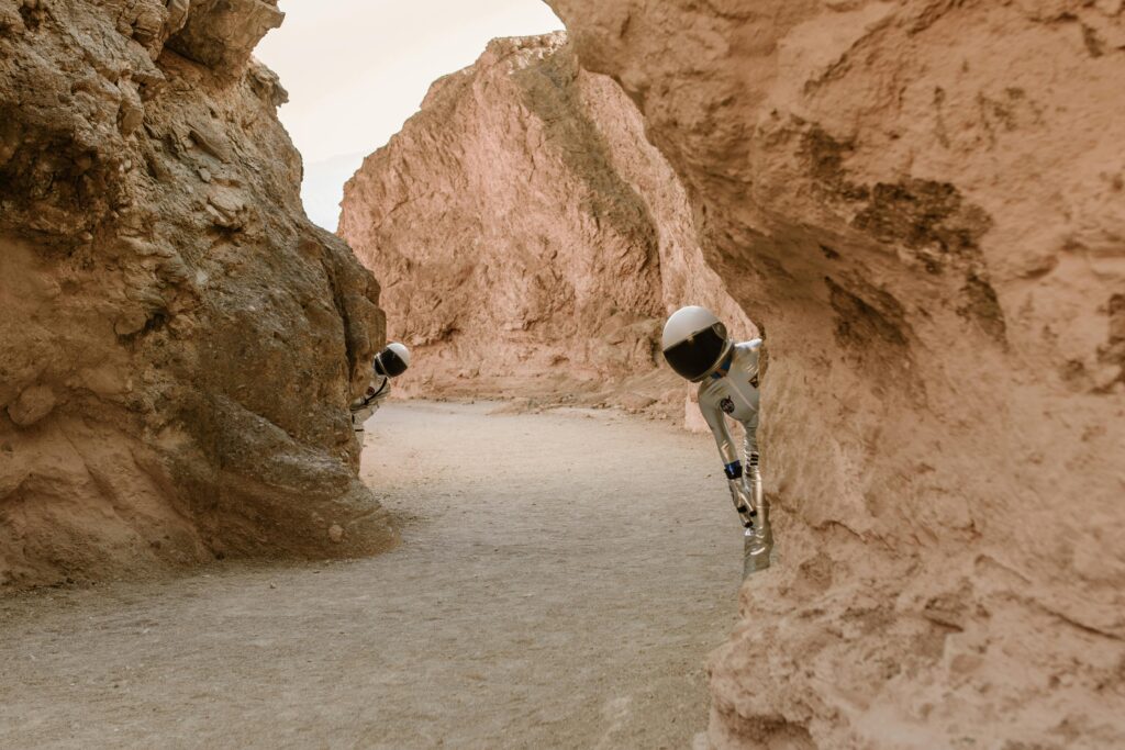 Astronauts peek from behind rocks in a fantasy extraterrestrial landscape.