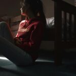 Contemplative young woman in hoodie sitting on the floor in dimly lit room, symbolizing solitude and introspection.