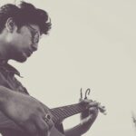 A black and white side view portrait of a male guitarist playing solo outdoors.