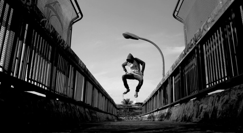 A man in a hoodie jumps lively on an urban bridge in black and white.