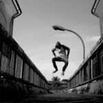 A man in a hoodie jumps lively on an urban bridge in black and white.