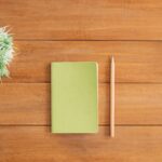 Flat lay of green notebook and pencil on wooden desk with plant. Perfect for creativity and organization themes.